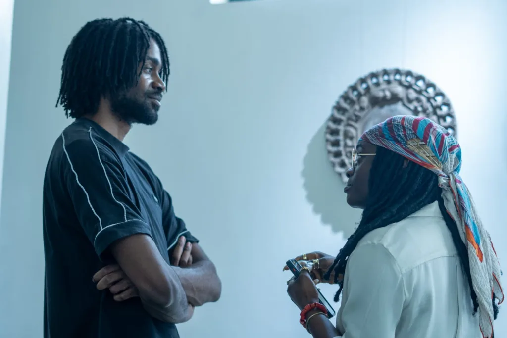 A man with dread on black shirt, his hands folded while listening to a lady on a white shirt with a scarf. They seem to be in a conversation.