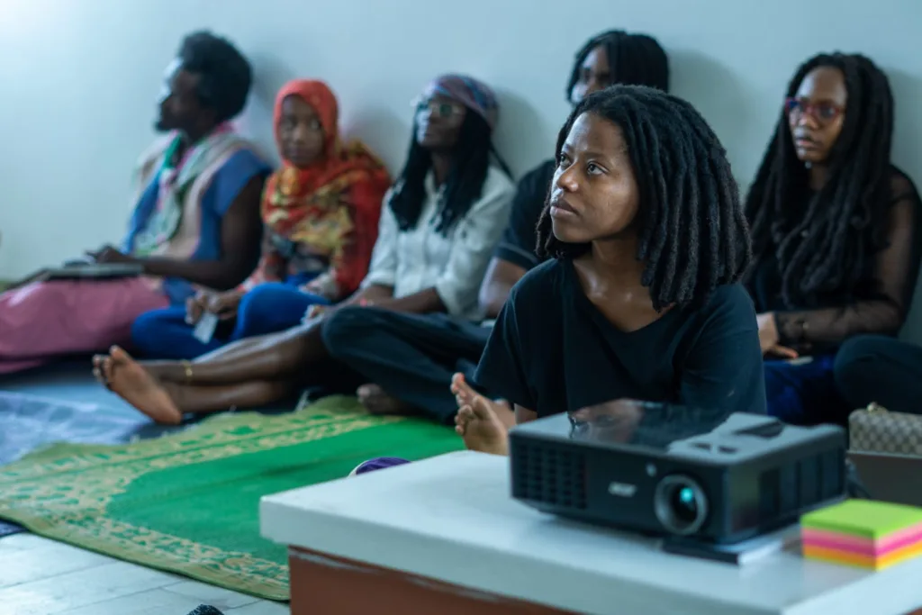 A picture of  Jubilian Ngaruwa wearing a black shirt and the audience, they are all seated on a mat and watching the Ancestry documentary.