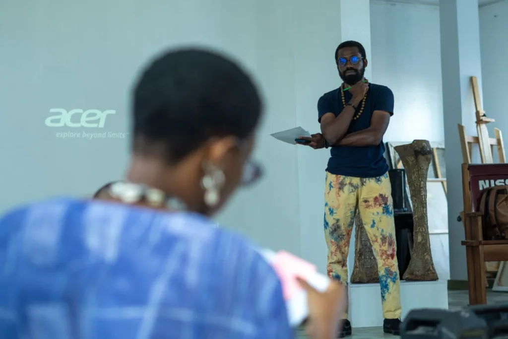 A picture of Patrick, DIDI museum's head curator standing while facilitating the Q&A, he's wearing a black top, wooded beads and an african print trousers. There's a lady sitting in front of him, but her picture is out of focus.